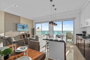 A modern living room with a sofa, a dining table, and a kitchen area with a view of the sea.