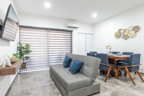 Un salon moderne avec un canapé gris, des coussins bleus, une table à manger en bois avec des chaises bleues, un téléviseur fixé au mur et un élément décoratif mural.