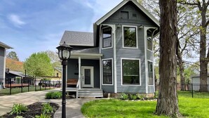Una casa a due piani con un portico, un giardino anteriore con erba e un albero, e una recinzione nera.