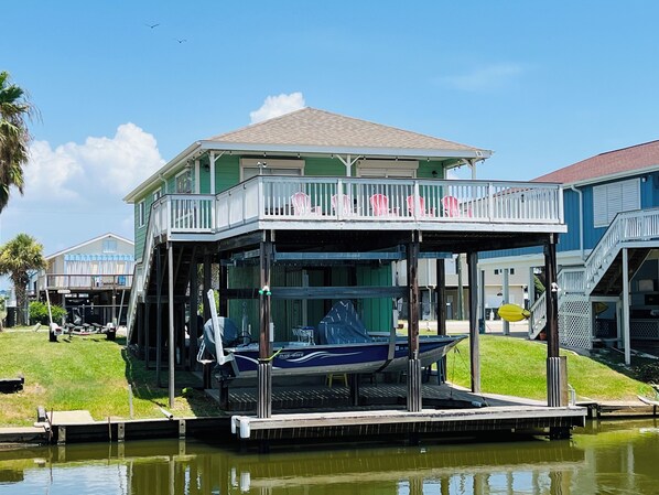 A waterfront building with a covered deck, a boat docked in front, and a grassy area with a palm tree.