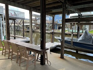 A covered dock area with wooden chairs and a table, overlooking a body of water with boats and houses in the background.
