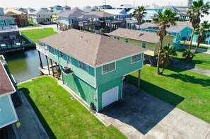A two-story house with a dock extending into a body of water, surrounded by other houses and palm trees.