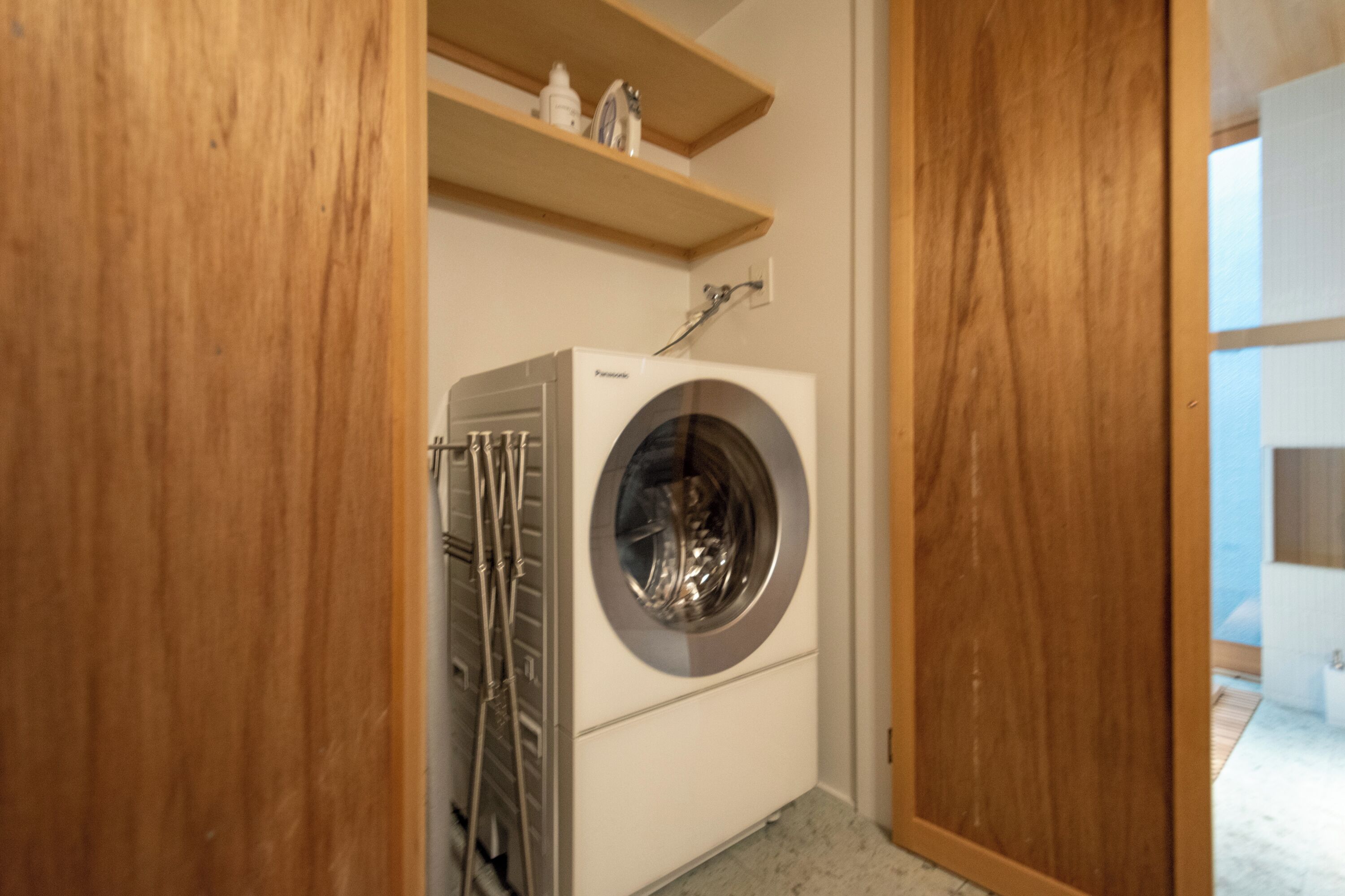 A compact laundry room with a front-loading washing machine, a drying rack, and wooden shelving above.