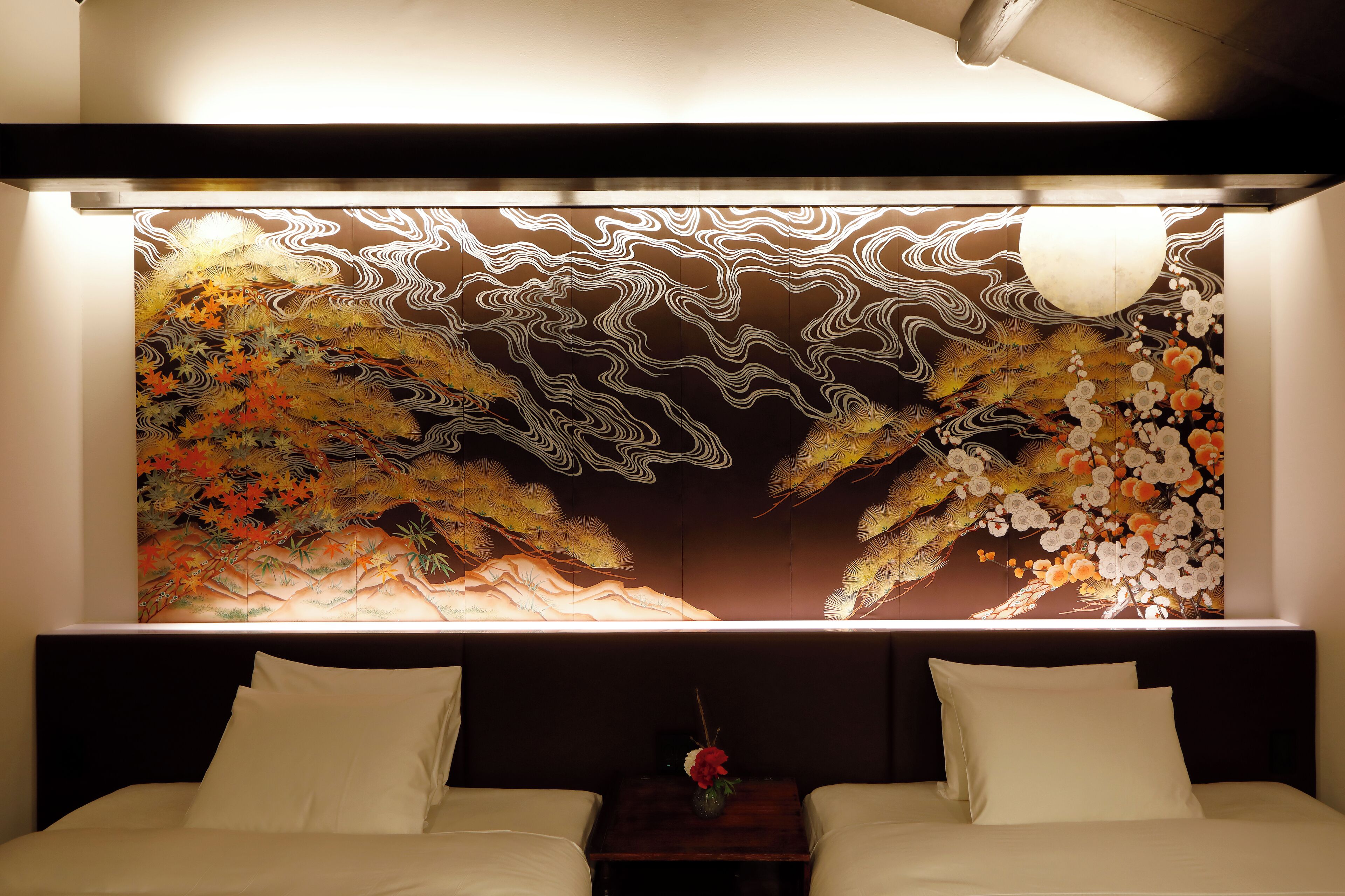 Habitación de hotel con un mural grande en la pared, una cama con ropa blanca y una mesita de noche con un jarrón de flores.