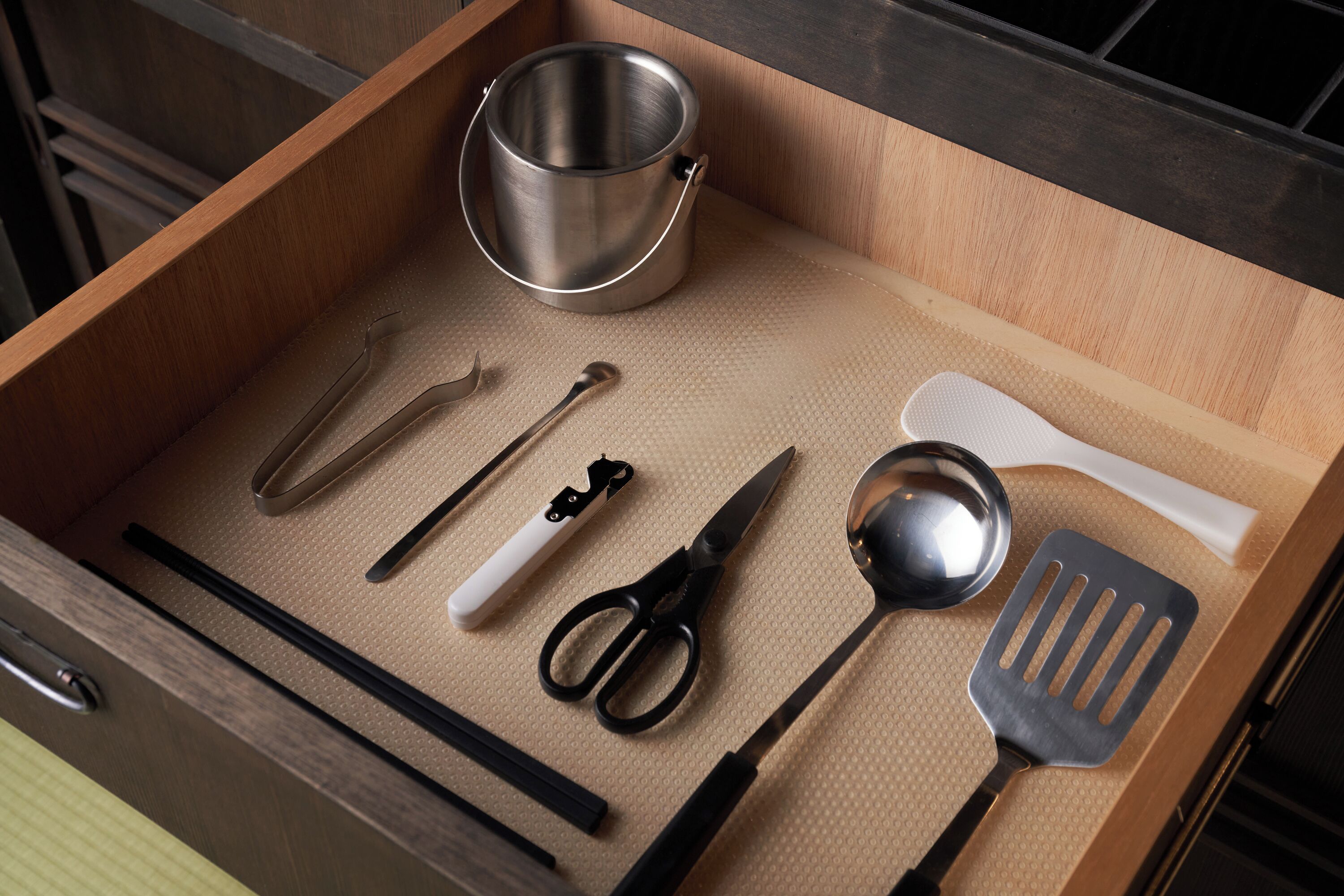 A kitchen drawer with a stainless steel pot, a white spatula, a pair of scissors, a black spatula, a black knife, and a black whisk.