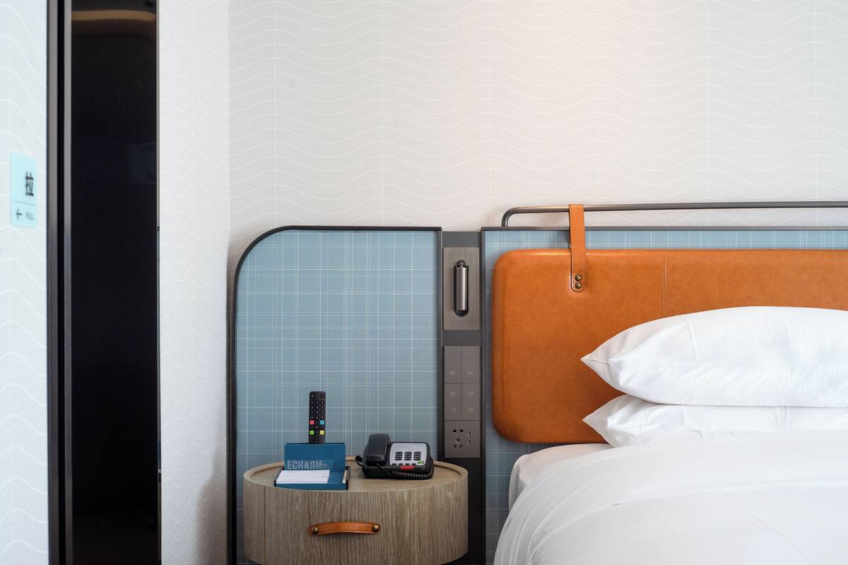 Habitación de hotel con cama, mesita de noche con teléfono y control remoto, y panel de pared con pomo.