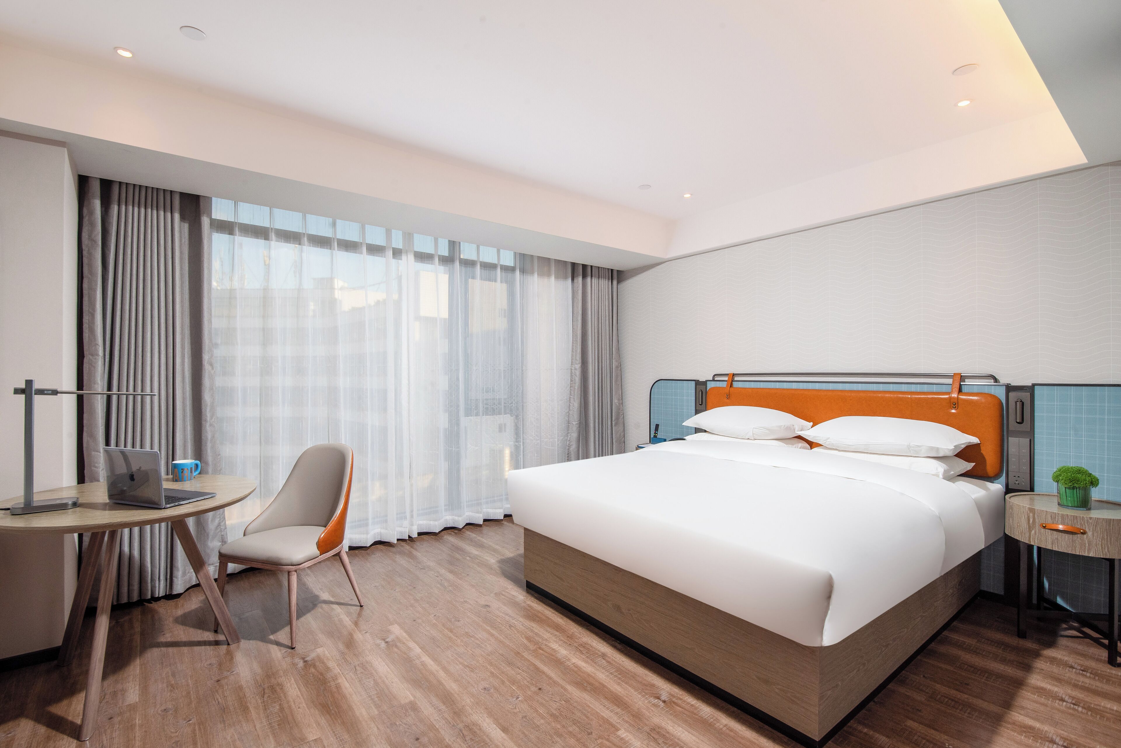 A modern hotel room with a large bed, a desk with a laptop, a chair, and a small table with a plant.