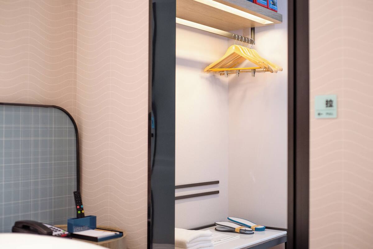 Habitación de hotel con cama, mesita de noche con control remoto, toalla y ropa colgada en un perchero.