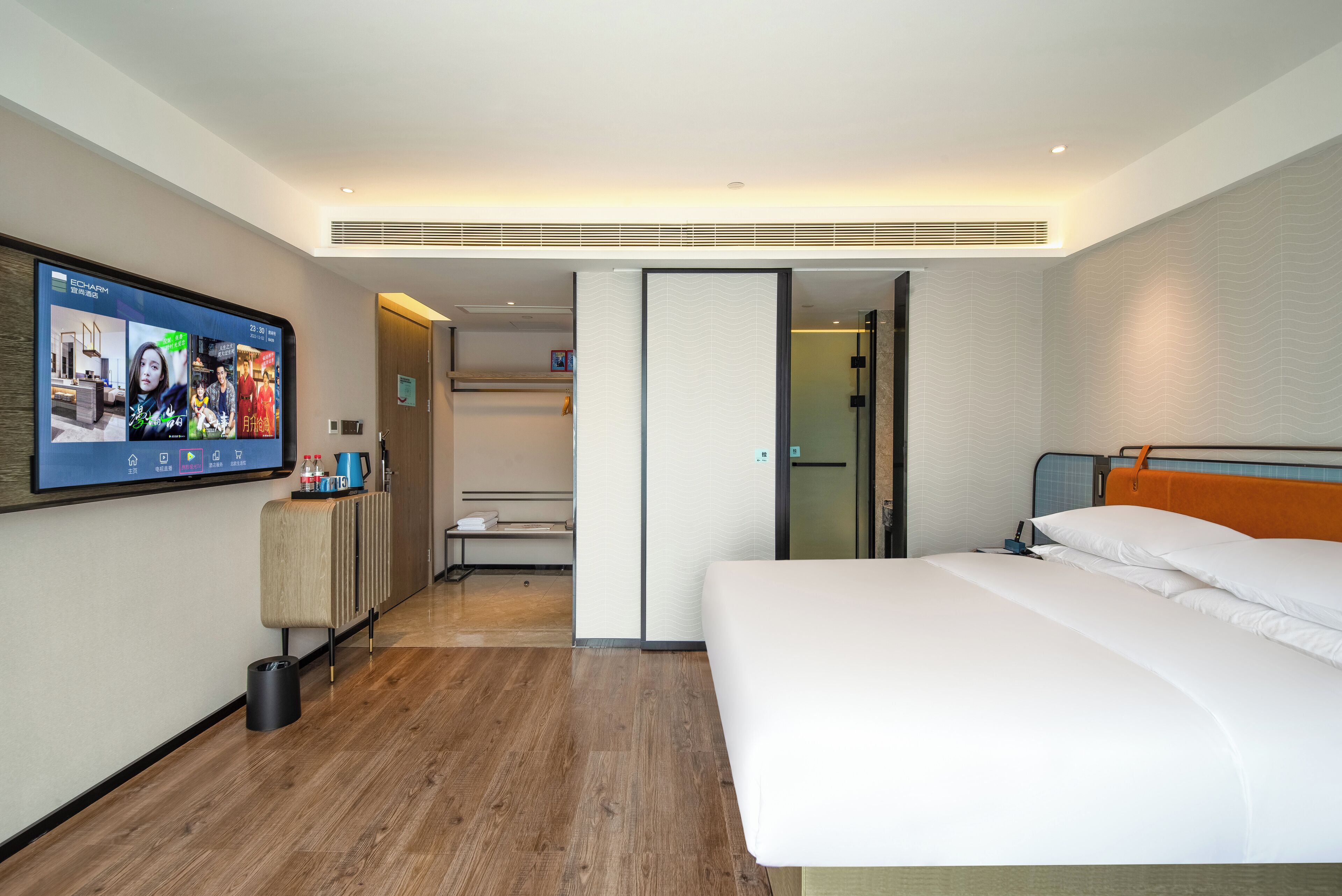 Habitación de hotel con un televisor de pantalla plana grande montado en la pared, una cama con ropa de cama blanca, piso de madera y un mueble pequeño con artículos de aseo.