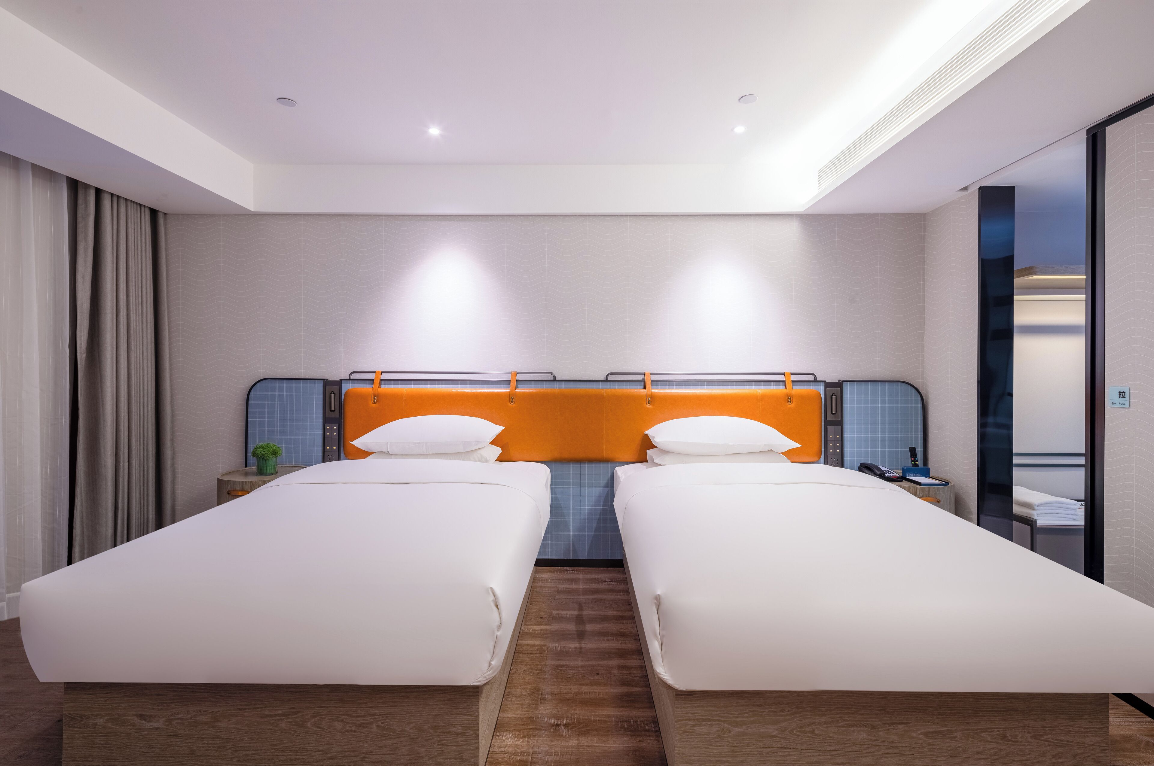 A hotel room with two single beds, a wooden floor, a modern orange headboard, and a small bedside table with a lamp.