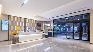 Een moderne hotellobby met een receptiebalie, een digitaal scherm en een glazen deur naar het cafégedeelte.