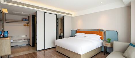 A modern hotel room with a large bed, a bedside table with a lamp, a small table with a plant, and a sofa.