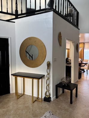 A staircase with black balusters, a round mirror with a gold frame, a small table with a black top, and a decorative vase on the floor.