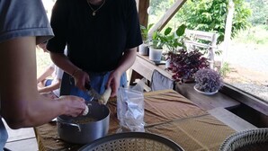 Un espace extérieur abrité, équipé d’une table servant de plan de travail pour la préparation des aliments, avec un panier rempli d’épis de maïs et une casserole posée sur la table.