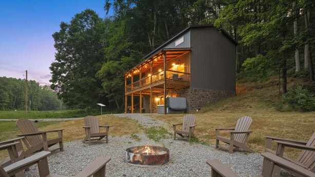 A rustic cabin with a stone foundation and a wooden deck, surrounded by a gravel area with Adirondack chairs. - Modern Rustic Retreat ~Newly Built with Hot Tub and EV charger, ~6mi to Ash Cave (Creola)