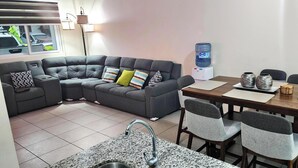 En moderne stue med en grå modulsofa, et træspisebord med fire stole, og et køkkenområde med en granitbordplade og vask.