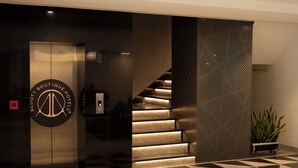 A modern hotel lobby with a geometric patterned floor, a staircase with illuminated steps, and a logo on the elevator door. - The Aldott Hotel (Gurugram)