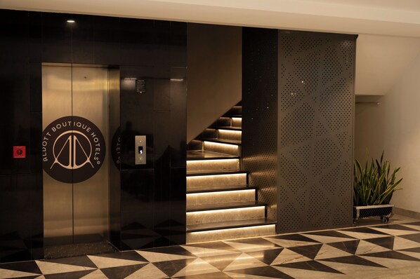 A modern hotel lobby with a geometric patterned floor, a staircase with illuminated steps, and a logo on the elevator door.