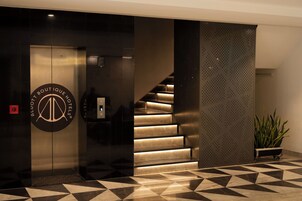 A modern hotel lobby with a geometric patterned floor, a staircase with illuminated steps, and a logo on the elevator door.