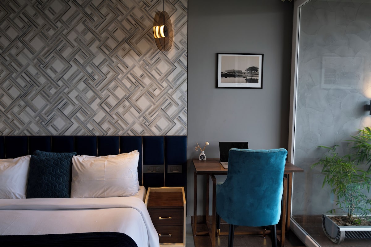 A hotel room with a patterned wall, a bed with white and dark blue pillows, a desk with a laptop, a blue chair, and a small plant.