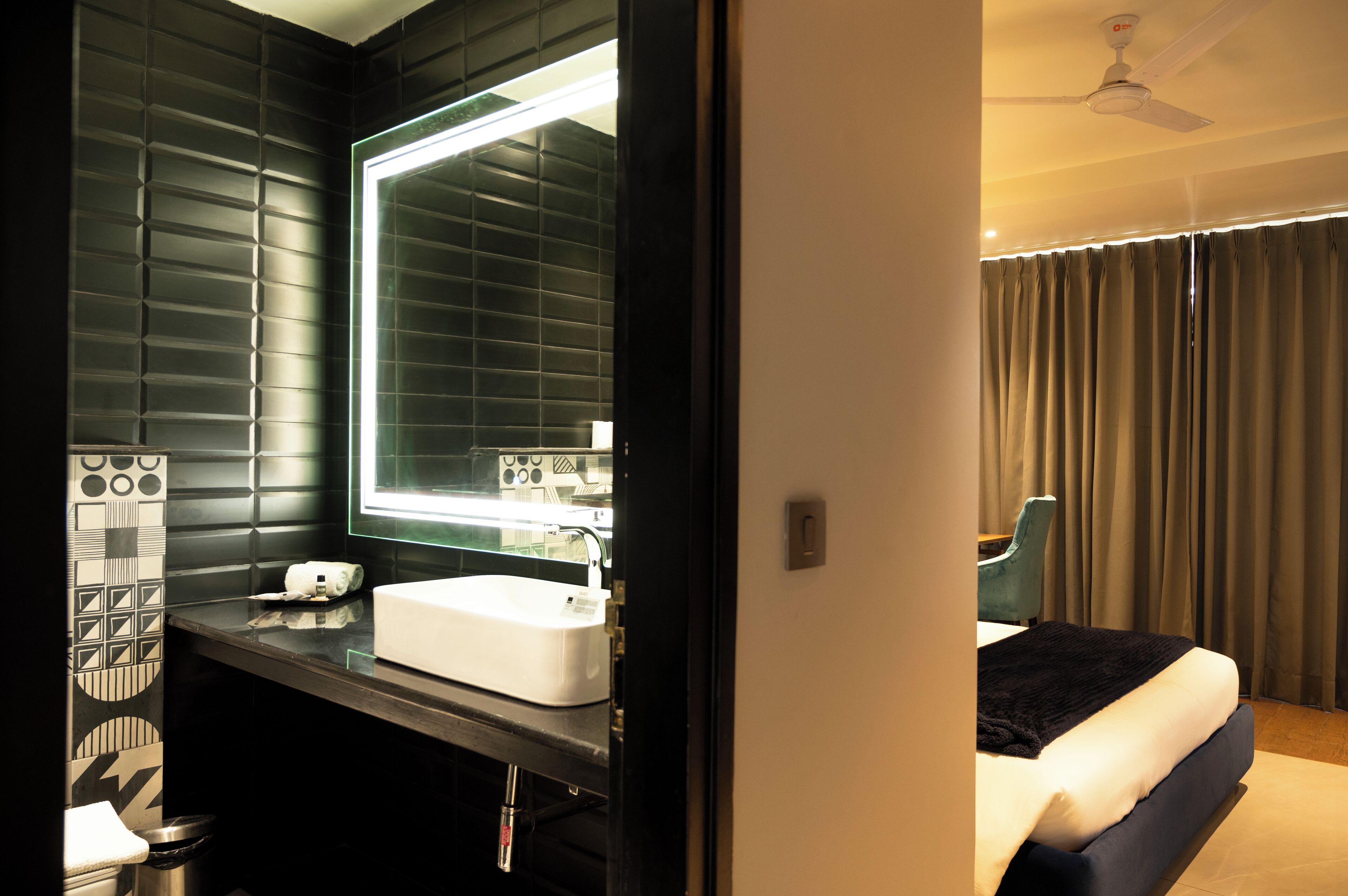 Ein modernes Hotelzimmer mit einem großen Spiegel, einem weißen Waschbecken, einem Bett mit dunklem Kopfteil und einem Deckenventilator.