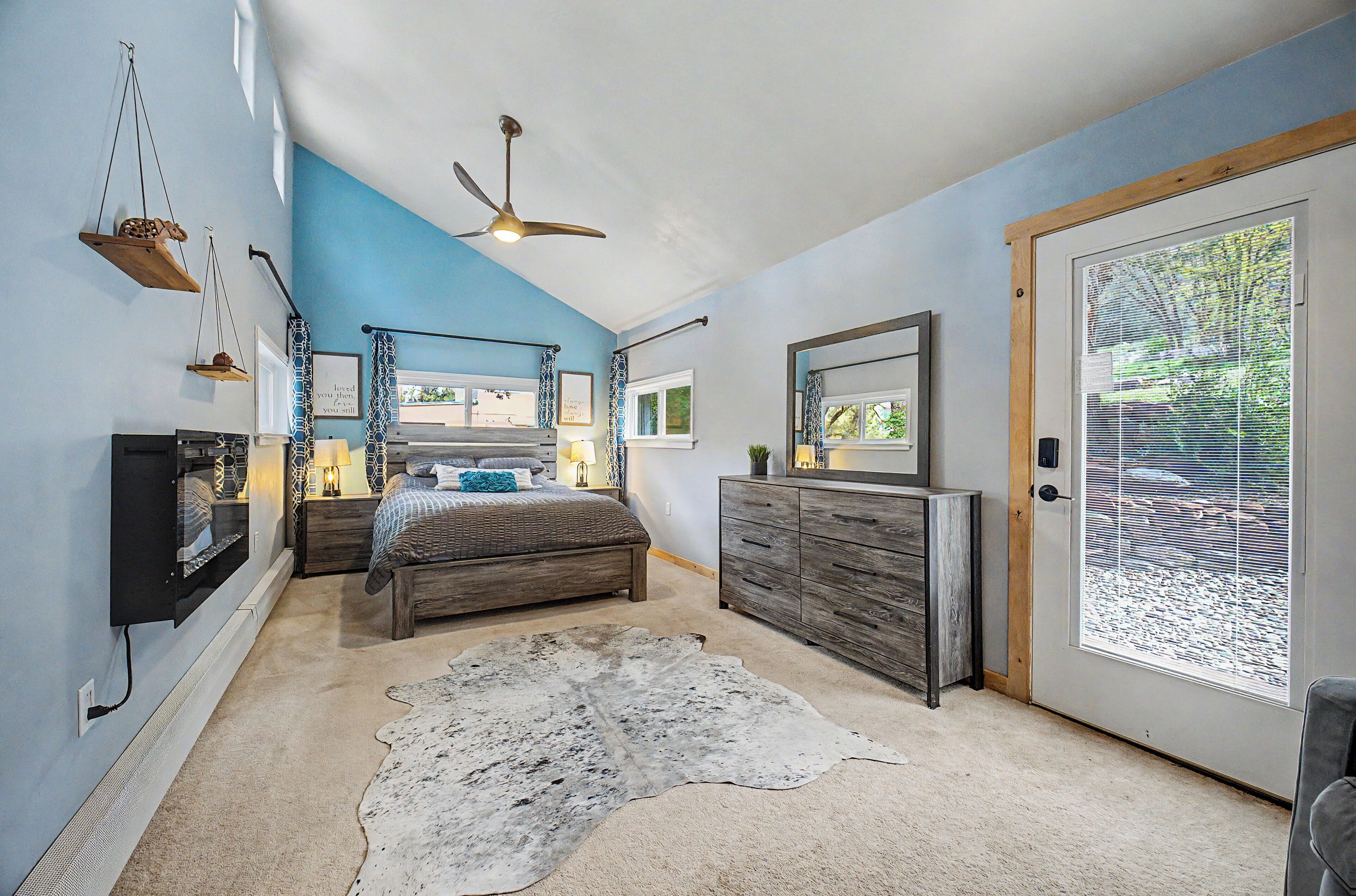 Une chambre à coucher comprenant un lit, une commode, un ventilateur de plafond et une grande fenêtre avec des stores.
