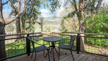 Une terrasse en bois avec une table ronde et des chaises, entourée d’une rambarde en métal, offrant une vue sur un paysage verdoyant, avec des arbres et, au loin, une chaîne de montagnes.