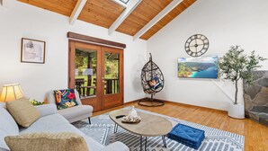 Uma sala de estar moderna com teto alto, vigas de madeira, um sofá, uma mesa de centro, uma poltrona, um vaso com planta e um quadro.