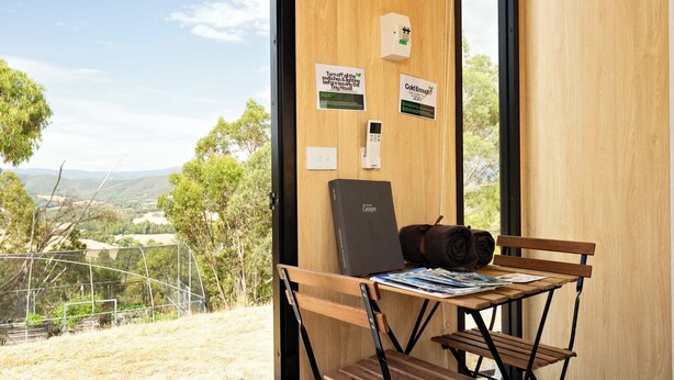 Une pièce avec une table en bois, une chaise et un sac à dos. Il y a des affiches au mur et une vue sur l’extérieur.