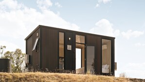 Une cabane moderne aux couleurs sombres, dotée d’une grande ouverture qui donne sur une chambre à coucher avec un lit et une fenêtre.