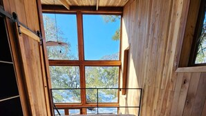 A wooden room with a large window, a bed with a beige blanket, and a wooden ceiling.