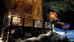 A modern wooden cabin with large windows and a balcony, surrounded by trees and snow.