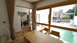 A room with a wooden dining table and chairs, a staircase, and large windows overlooking a street. - RIM HuaHin Villa By RoomQuest (Hua Hin)