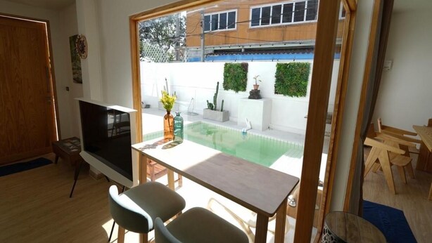 A room with a TV, a dining table with chairs, a small pool, and a view of a building with windows. - RIM HuaHin Villa By RoomQuest (Hua Hin)