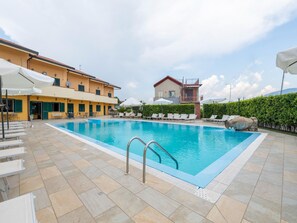 A swimming pool with sun loungers and umbrellas, surrounded by a tiled patio and a building with a yellow facade.