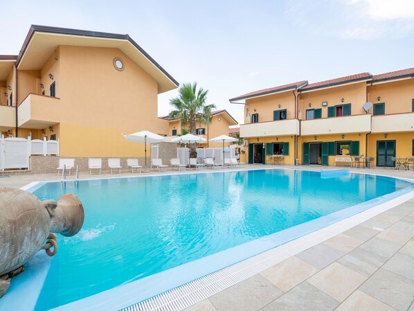 A swimming pool with lounge chairs and umbrellas, surrounded by yellow buildings with balconies and windows.