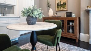 Une cuisine moderne avec des meubles blancs, une table ronde en marbre, des chaises recouvertes d’un tissu vert, une plante en pot et un tableau accroché au mur.