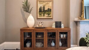 Un meuble en bois avec des portes en verre, un vase blanc, un tableau et un miroir.