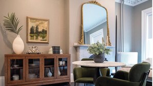 Une pièce comprenant une table ronde, deux chaises vertes, un meuble en bois doté de portes en verre, un grand miroir, un tableau représentant un paysage encadré, un vase blanc et une plante en pot.