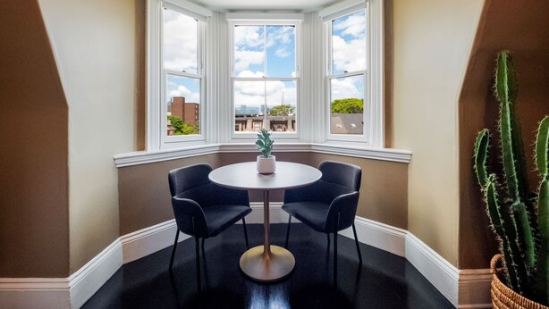 A corner room with a bay window, a round table with two chairs, a potted plant, and a cactus on the floor. - The Chester: Modern, Cool, Heart Of South End (Boston)