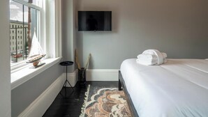 A bedroom with a bed, a TV mounted on the wall, a window with a view of buildings, and a floor lamp. - The Chester: Modern Luxury, Historic Charm (Boston)