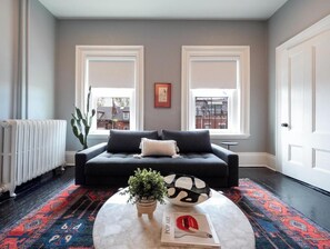 Una sala de estar con un sofá negro, una mesa redonda de centro con un libro y una planta, y dos ventanas con marcos blancos.