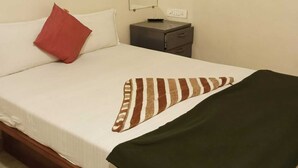Een enkel bed met een witte matras, een rood kussen, een gestreept dekbed en een groene plaid. Er staat een nachtkastje met een lamp en een stopcontact.
