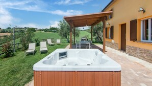 Un jacuzzi bajo una pérgola de madera, un patio con sillas y vistas a un paisaje verde.