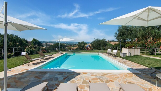Una piscina rectangular con losetas de piedra, rodeada de sillas de descanso y sombrillas, con un fondo de vegetación y colinas al lejano.