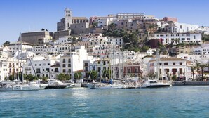 A coastal town with a historic castle on a hill, a marina with sailboats, and white buildings with balconies.