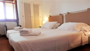 A hotel room with a large bed, two bedside tables, a window with blinds, and a door with a shutter.