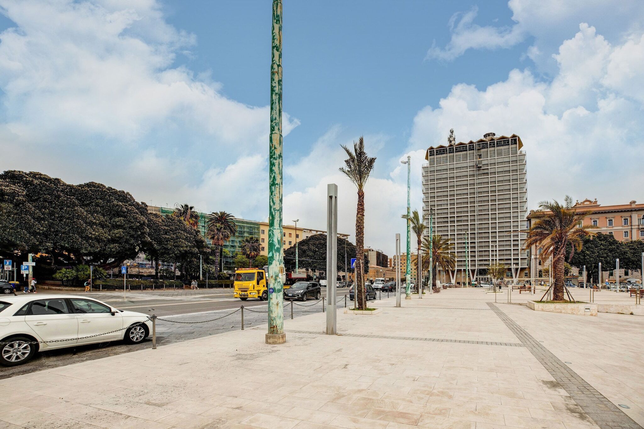 A city street with a white car parked on the side, a yellow truck, and a tall building in the background.