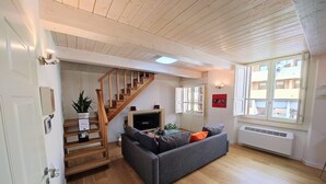 A living room with a grey sofa, wooden staircase, TV, and a large window with blinds. - Civico36 Feels Like Home (Cagliari)