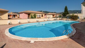 Ein Swimmingpool mit klarem, blauem Wasser, umgeben von einer gefliesten Terrasse, neben Gebäuden im mediterranen Baustil.